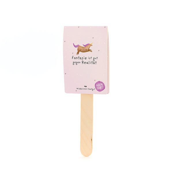 Holzstab mit rosa Karte, darauf ein gezeichnetes Einhorn und der Text "Fantasie ist gut gegen RealitÀt", darunter steht "Wildblumen-Saatgut".