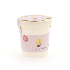 Pflanzbecher mit rosa Etikett, auf dem "Schokoladenblume fĂŒr dich" und eine Zeichnung einer Blume ĂŒber einem SchokoladenstĂŒck zu sehen ist.