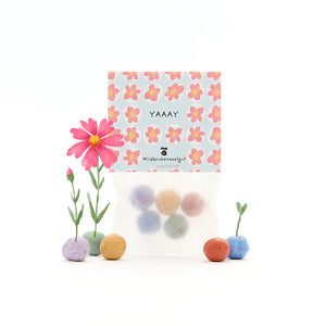 Tüte mit bunten Wildblumensamen-Kugeln, Blumenmuster auf der Verpackung und kleinen, blühenden Blumen daneben.