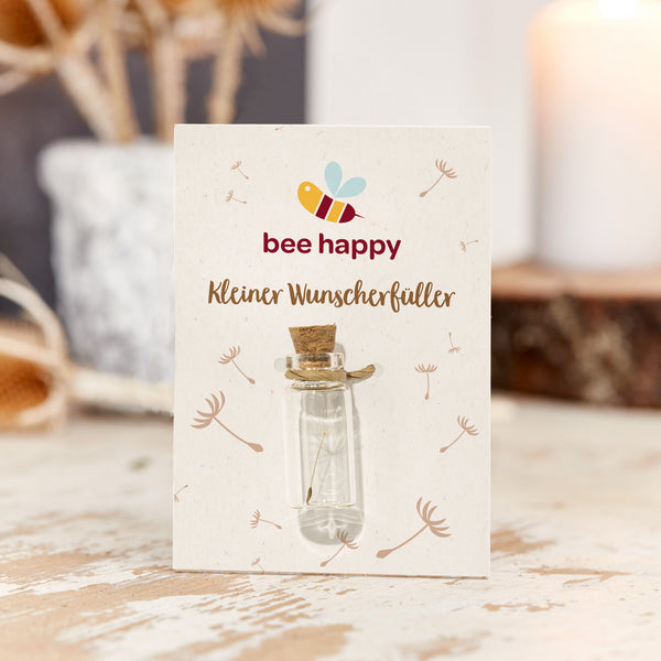 Kleine Glasflasche mit Korken auf einer Karte mit der Aufschrift „bee happy Kleiner Wunscherfüller“, dekoriert mit Pusteblumenmotiven.