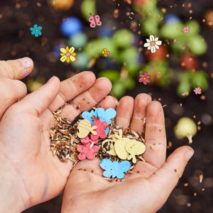 Zwei schmutzige Kinderhände halten Blumensamen und bunte Papierformen in Form von Blumen und Schmetterlingen über Erde.