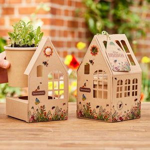 Zwei dekorative, kartonierte Häuschen mit Blumen- und Insektenmotiven, eines mit einem Pflanztopf mit grünen Keimlingen, beide auf einem Holztisch im Garten platziert.