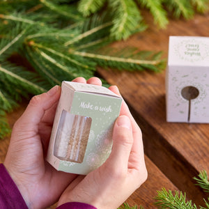 Hände halten eine kleine Geschenkbox mit der Aufschrift "Make a wish" neben Tannenzweigen auf einem Holztisch.