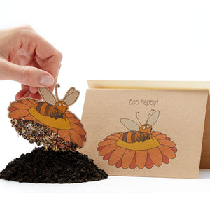 Eine Hand, die eine Aussaatkarte in Form einer Biene hochhält, mit dem Slogan "Bee happy!" darauf und einem Haufen Erde darunter.
