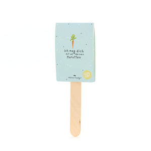Ein Holzstäbchen mit hellblauem Schild, darauf eine Karottenzeichnung und der Text „Ich mag dich mit all deinen Karotten“.