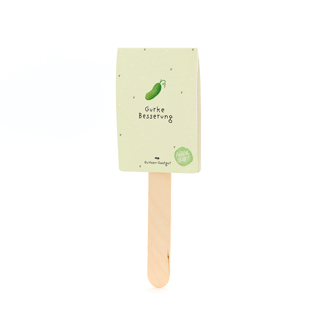 Ein Holzstab mit einem beige-grünen Papieretikett, auf dem eine gezeichnete Gurke und der Text "Gurke Besserung" zu sehen sind.