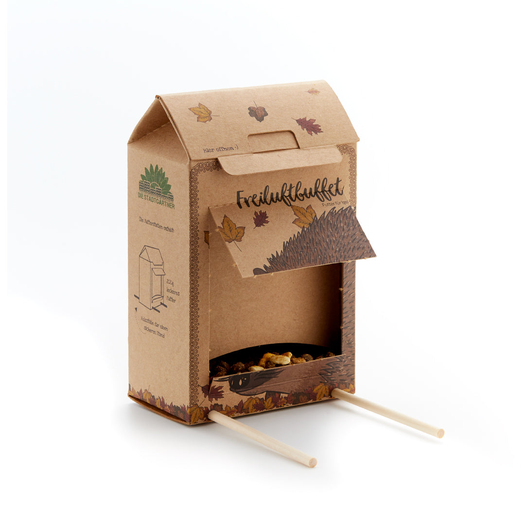 Pappkarton mit geöffneter Klappe, Herbstblättern, Igel-Illustration und zwei Holzstäben, dient als Futterstation für Igel.