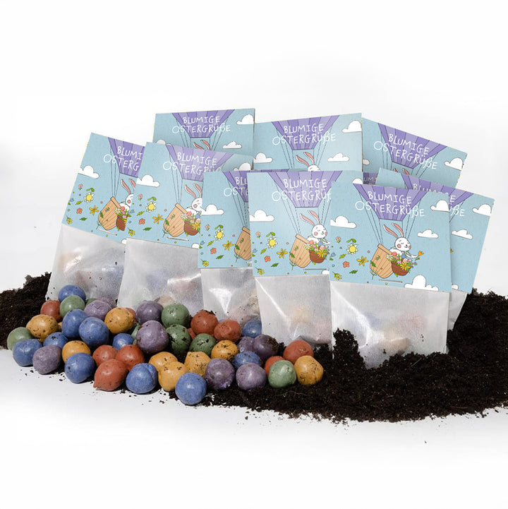 Mehrere Tütchen mit der Aufschrift "Blumige Ostergrüße" und einem Osterhasenmotiv, davor bunte Samenbomben auf Erde.