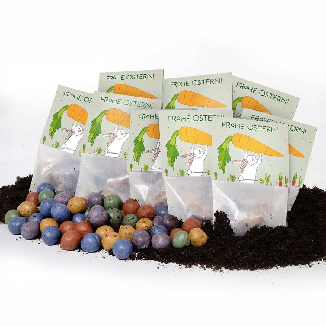 Mehrere Ostern-Samentütchen mit der Aufschrift "Frohe Ostern!" und bunter Samen-Konfetti auf Erde.