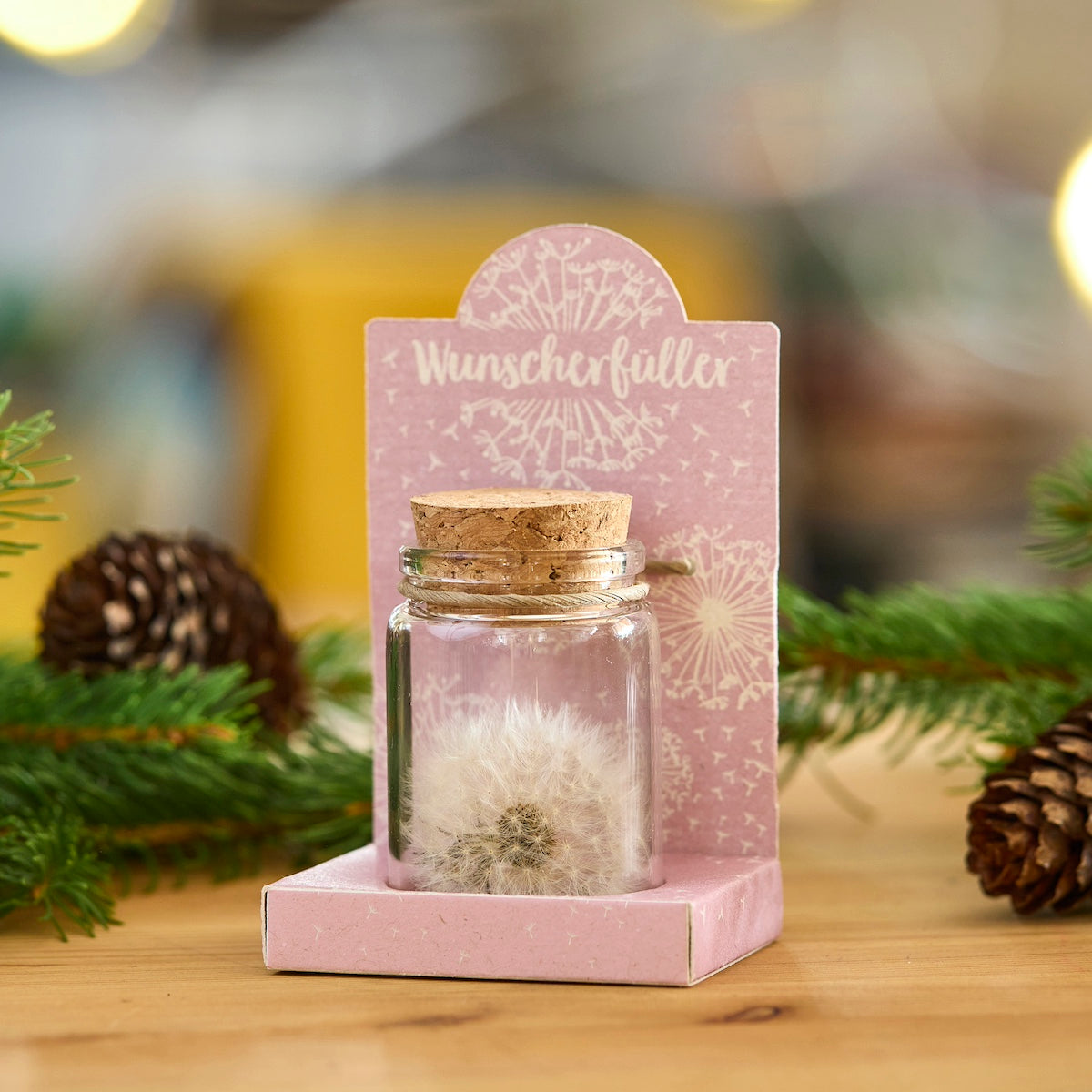 Kleines Glas mit Korken, enthält eine Pusteblume, steht auf einer rosa Verpackung mit der Aufschrift "Wunscherfüller", dekoriert mit Tannenzweigen und Zapfen.