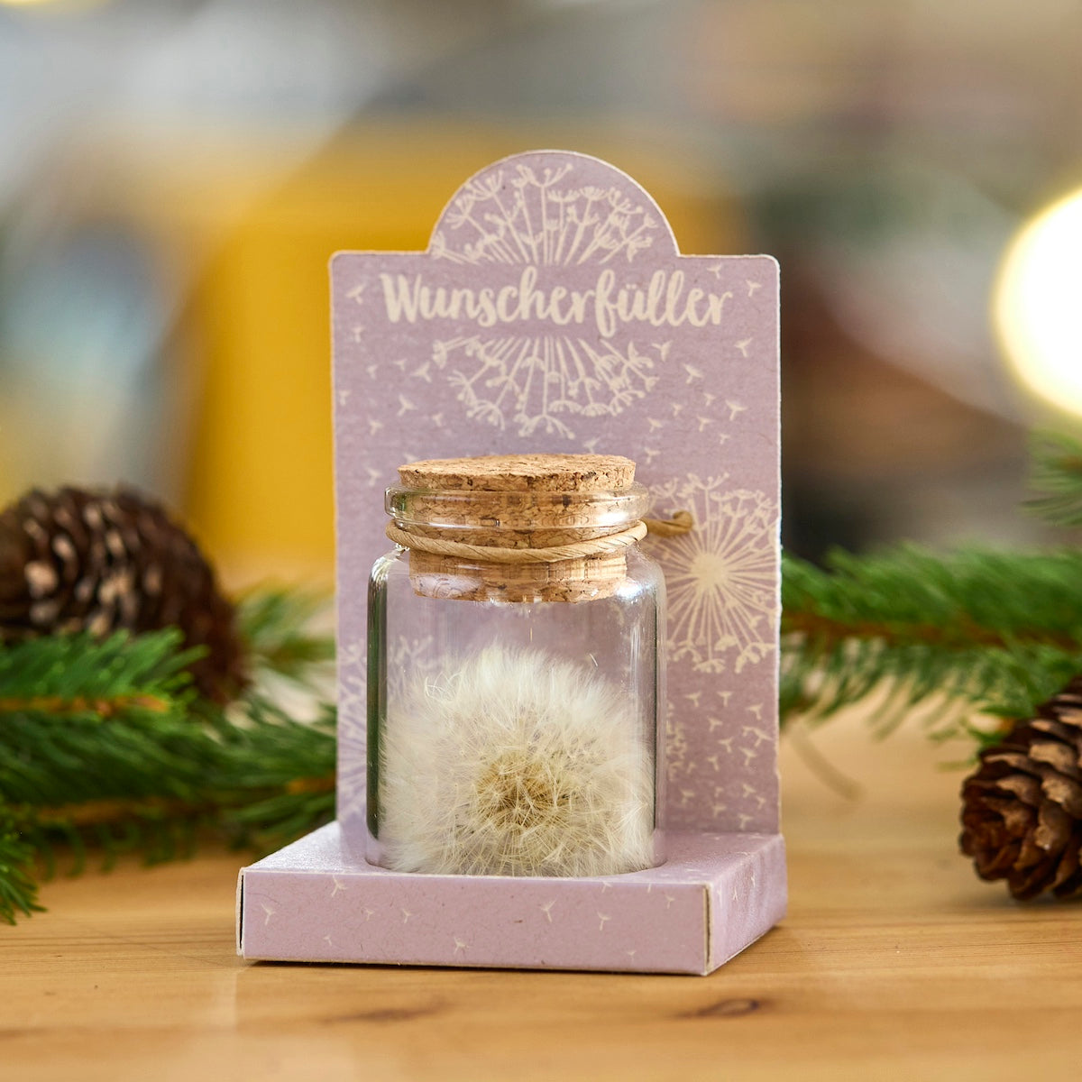 Glasfläschchen mit Pusteblume und Korken, auf einer lilafarbenen Verpackung mit der Aufschrift "Wunscherfüller", dekoriert mit Tannenzweigen und Zapfen.