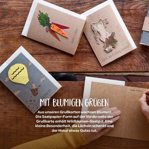 Verschiedene Grußkarten aus braunem Recyclingpapier mit bunten Saatpapier-Motiven, auf einem Holztisch liegend.