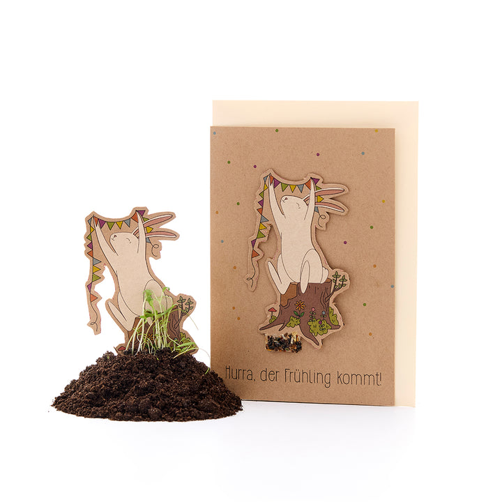 Eine Frühlingsgrußkarte mit einem Hasenmotiv und der Aufschrift „Hurra, der Frühling kommt!“, daneben ein ausgeschnittener Hase mit Keimlingen in Erde.
