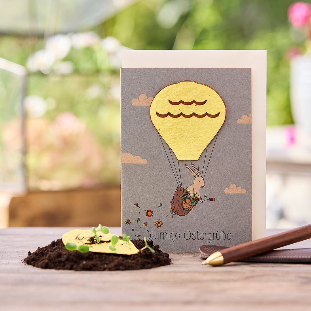 Osterkarte mit Hase im Heißluftballon, daneben Erde mit keimenden Pflänzchen und ein Stift.