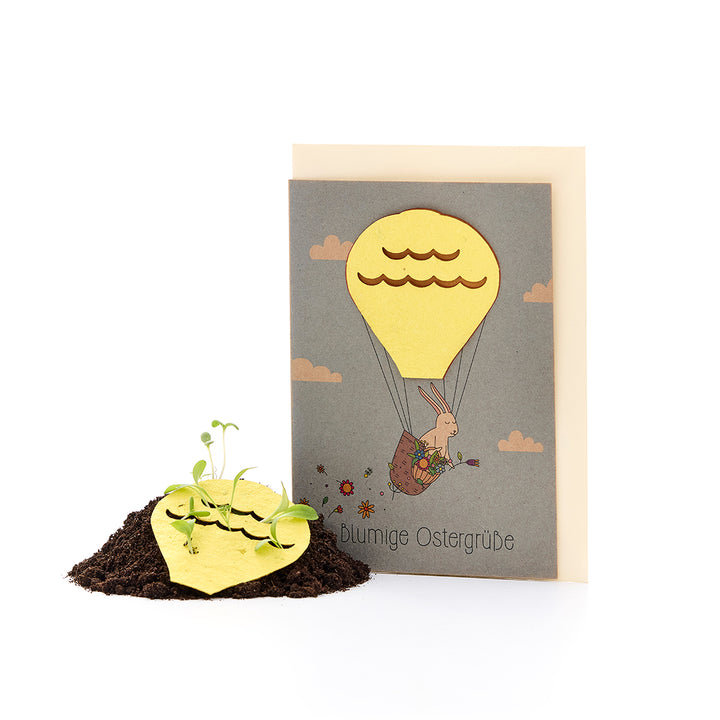 Osterkarte mit einem Hasen in einem Heißluftballon, dazu Samenpapier in Herzform, aus dem kleine Pflanzen in Erde wachsen.