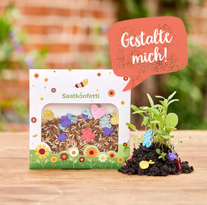 Packung mit Saatkonfetti und kleinen, bunten Papierfiguren, daneben junge Pflanzen, auf Holztisch, im Hintergrund unscharfe Natur.