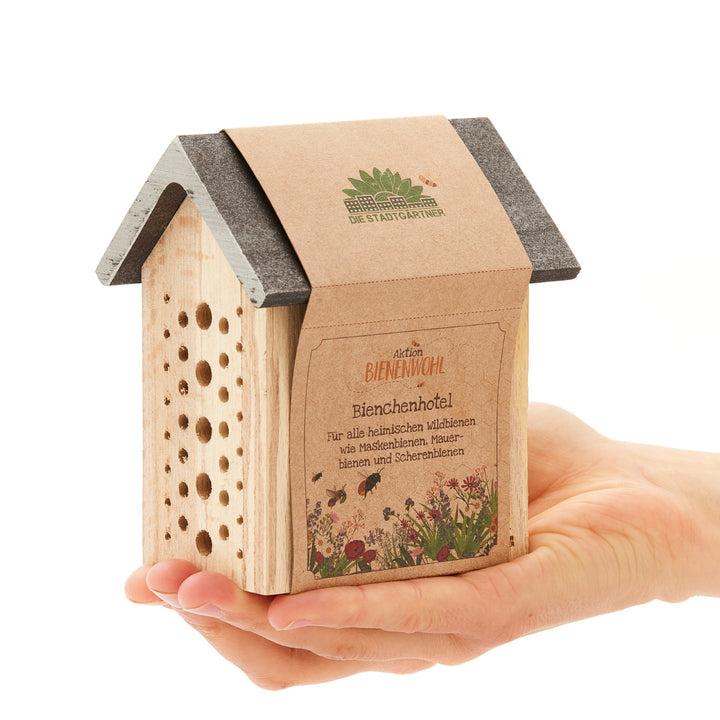 Eine Hand hält ein kleines Bienenhotel aus Holz, mit der Aufschrift "Aktion Bienenwohl Bienenhotel" auf Deutsch.