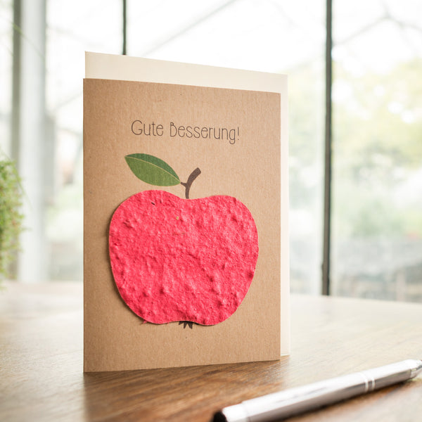 Grußkarte mit dem Text "Gute Besserung!" und einem roten Apfel auf einem Holztisch.