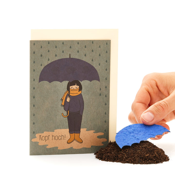 Hand platziert eine blaue Papierwelle auf einem Haufen Erde vor einer Grußkarte mit einer gezeichneten Figur unter einem Regenschirm und dem Text 'Kopf hoch!'"