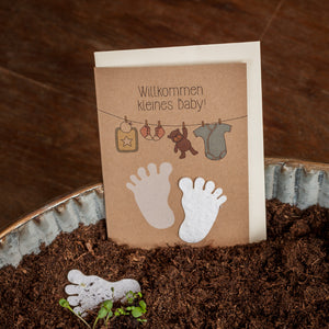 Glückwunschkarte zur Geburt mit der Aufschrift "Willkommen kleines Baby!" und Abbildungen von Babyfüßen, Kleiderbügel mit Strampler und Spielzeug, auf Erde und Holzhintergrund.