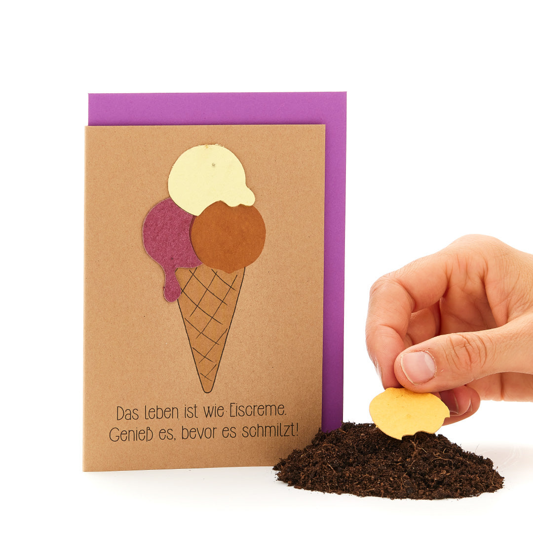 Hand, die ein herzförmiges Keks in Erdboden steckt, neben einer Grußkarte mit Eistüten-Motiv und dem Spruch 'Das Leben ist wie Eiscreme. Genieß es, bevor es schmilzt!'"