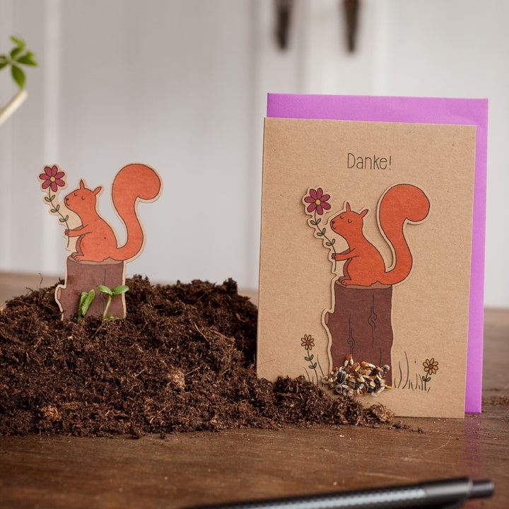 Grußkarte mit Eichhörnchen-Motiv und dem Wort "Danke!" darauf, neben einer ausgeschnittenen Eichhörnchen-Figur auf Erde.