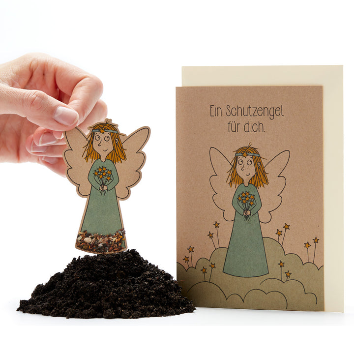 Hand hält eine Aussaatpapier-Figur eines Engels über Erde neben einer Karte mit Text 'Ein Schutzengel für dich'.