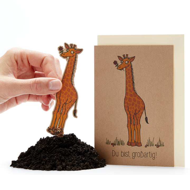 Eine Hand steckt eine Karte in Form einer Giraffe in Erde, daneben eine Grußkarte mit einer Giraffenzeichnung und dem Text "Du bist großartig!".