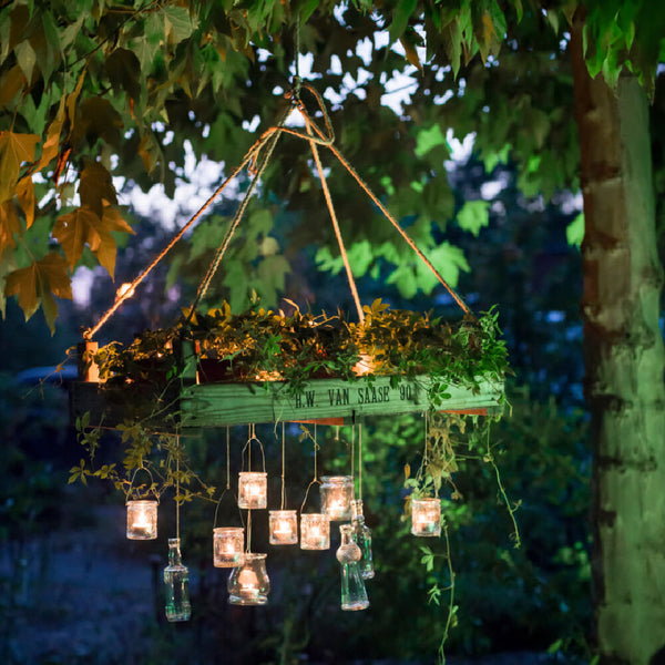 Hängende Blumenampel mit Pflanzen und Kerzen in Gläsern beleuchtet im Abendlicht