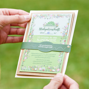 Zwei Hände halten eine Blühpatenschafts-Urkunde auf grünem Hintergrund, mit floraler Umrahmung und Text zum Verschenken einer Blumenwiese.